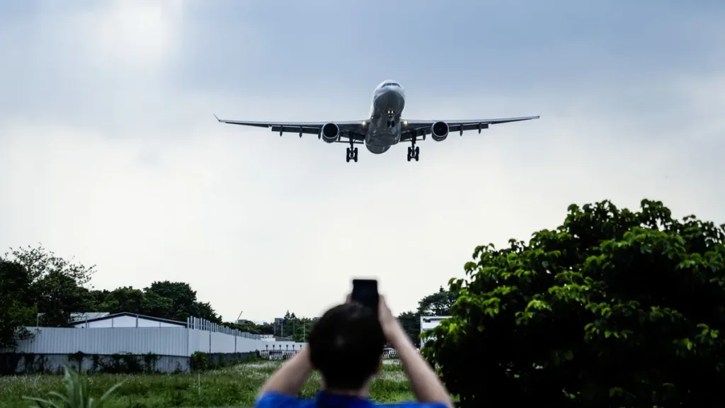 Passenger jet on final approach over trees while a person, seen from behind, photographs it on a smartphone