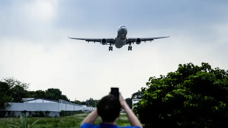 Passenger jet on final approach over trees while a person, seen from behind, photographs it on a smartphone