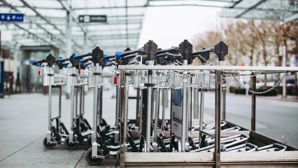 Row of empty luggage trolleys lined up outside an airport terminal building under a glass canopy
