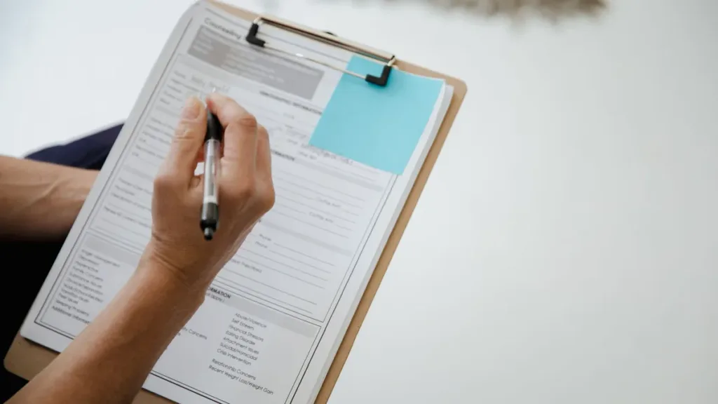 Close-up of a hand filling in a printed form on a wooden clipboard with a pen, a blue sticky note on the page