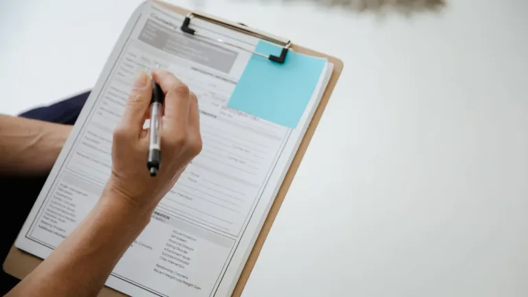 Close-up of a hand filling in a printed form on a wooden clipboard with a pen, a blue sticky note on the page