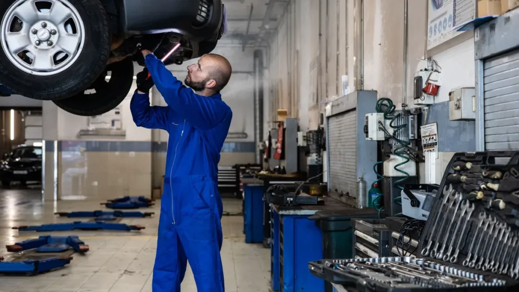 Mechanic in blue overalls inspecting the underside of a raised car in a workshop lined with tool cabinets