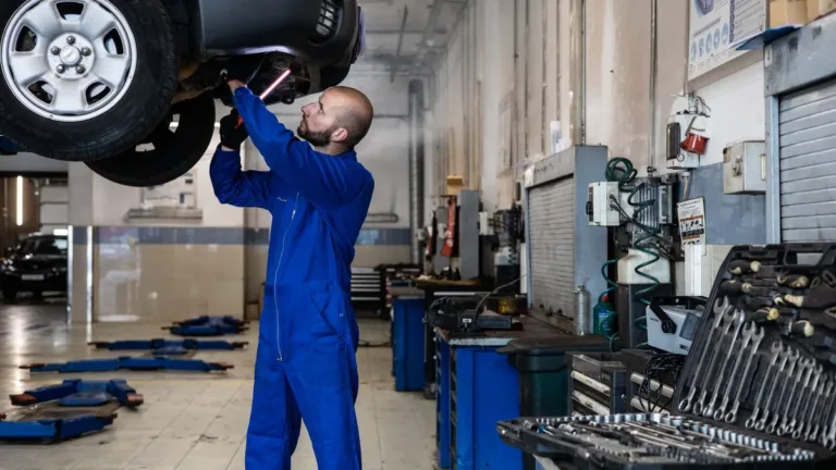 Mechanic in blue overalls inspecting the underside of a raised car in a workshop lined with tool cabinets