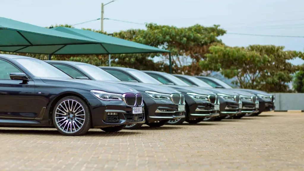 A row of dark executive saloon cars parked side by side on a paved forecourt under a green canopy