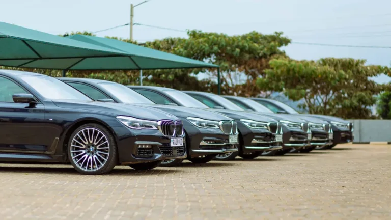 A row of dark executive saloon cars parked side by side on a paved forecourt under a green canopy