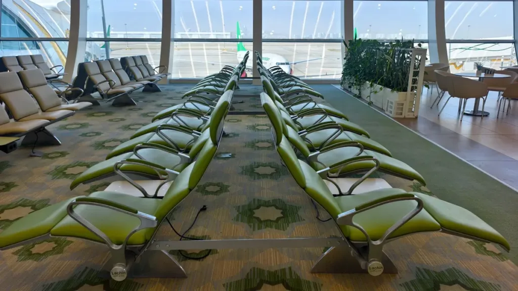 Empty airport waiting area with rows of green seats, patterned carpet and tall windows overlooking the apron