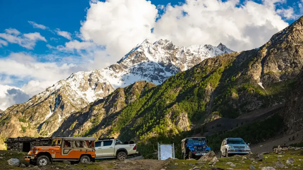 Jeeps and pickup trucks parked on a grassy ledge below snow-capped mountains and green slopes under a blue sky
