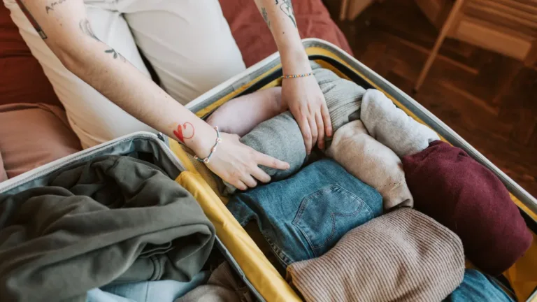 Hands folding jumpers, jeans and knitwear into an open silver suitcase while packing for a trip