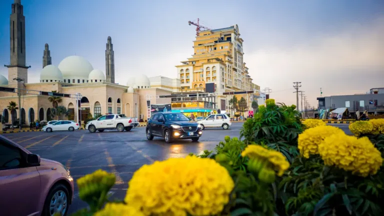 Cars driving along a wide city road in Wah, Pakistan, with a domed mosque and a tall hotel building behind marigolds