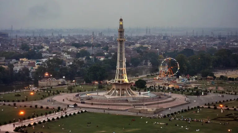Minar-e-Pakistan in Lahore at dusk, surrounded by lawns, walkways and a lit fairground wheel, the city beyond