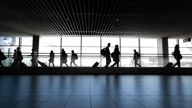 Silhouetted travellers with suitcases walking past tall windows along a bright airport terminal concourse