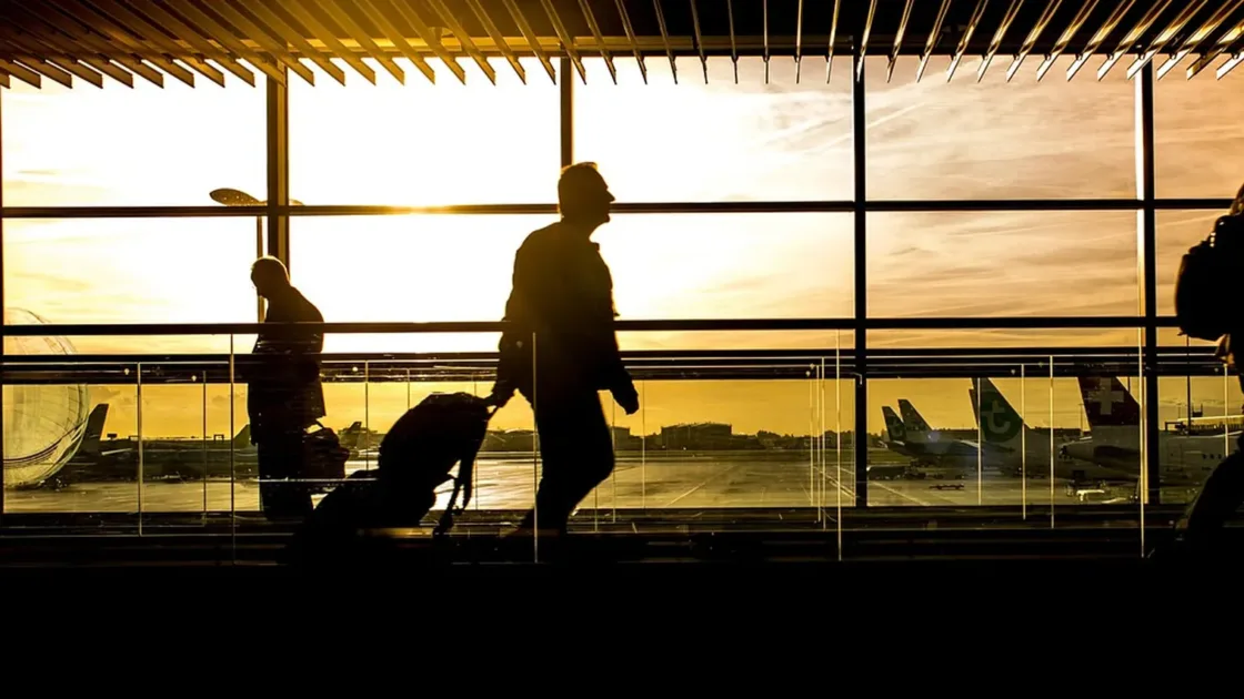 Silhouettes of travellers pulling luggage through an airport terminal with aircraft visible outside at sunset