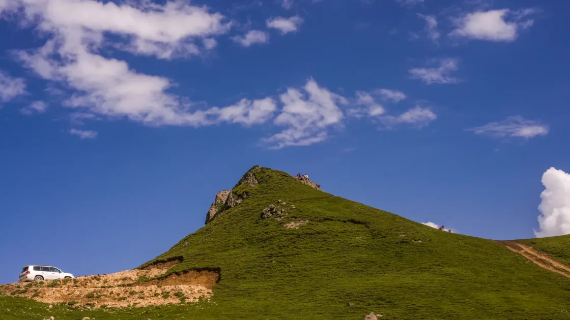 A white SUV parked on a grassy green hilltop in Khyber Pakhtunkhwa under a blue sky with white clouds