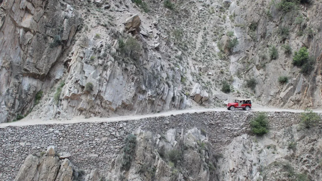 A red jeep travelling along a narrow road cut into a steep rocky cliff in the mountains near Skardu