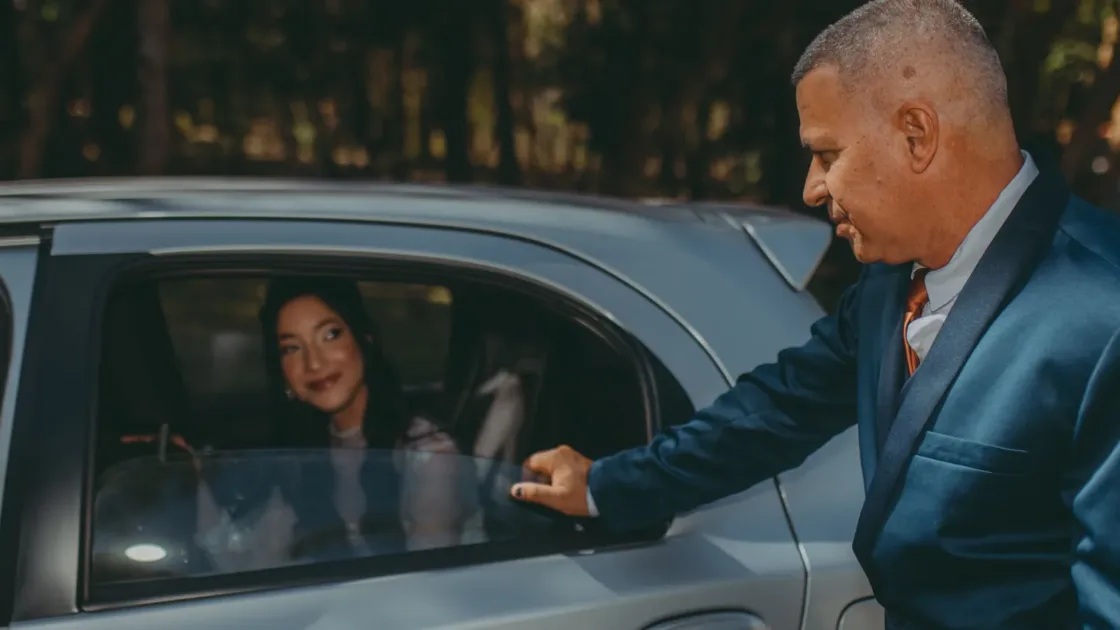 A driver in a blue suit opens the rear door of a silver car for a passenger seated inside