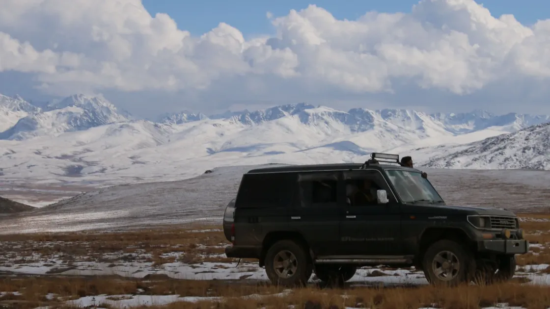 A dark 4x4 SUV driving across snow-dusted high ground in Gilgit-Baltistan with snowy peaks behind