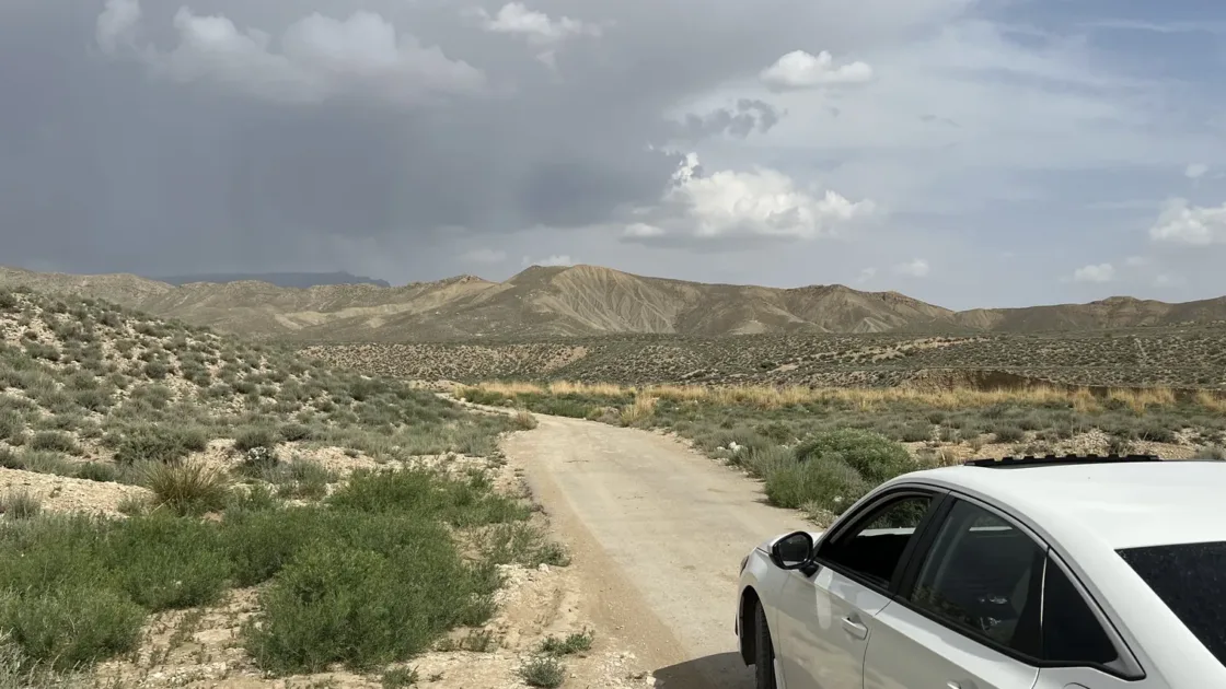 A white saloon car parked on a dirt road among dry scrub and low hills outside Quetta under a cloudy sky